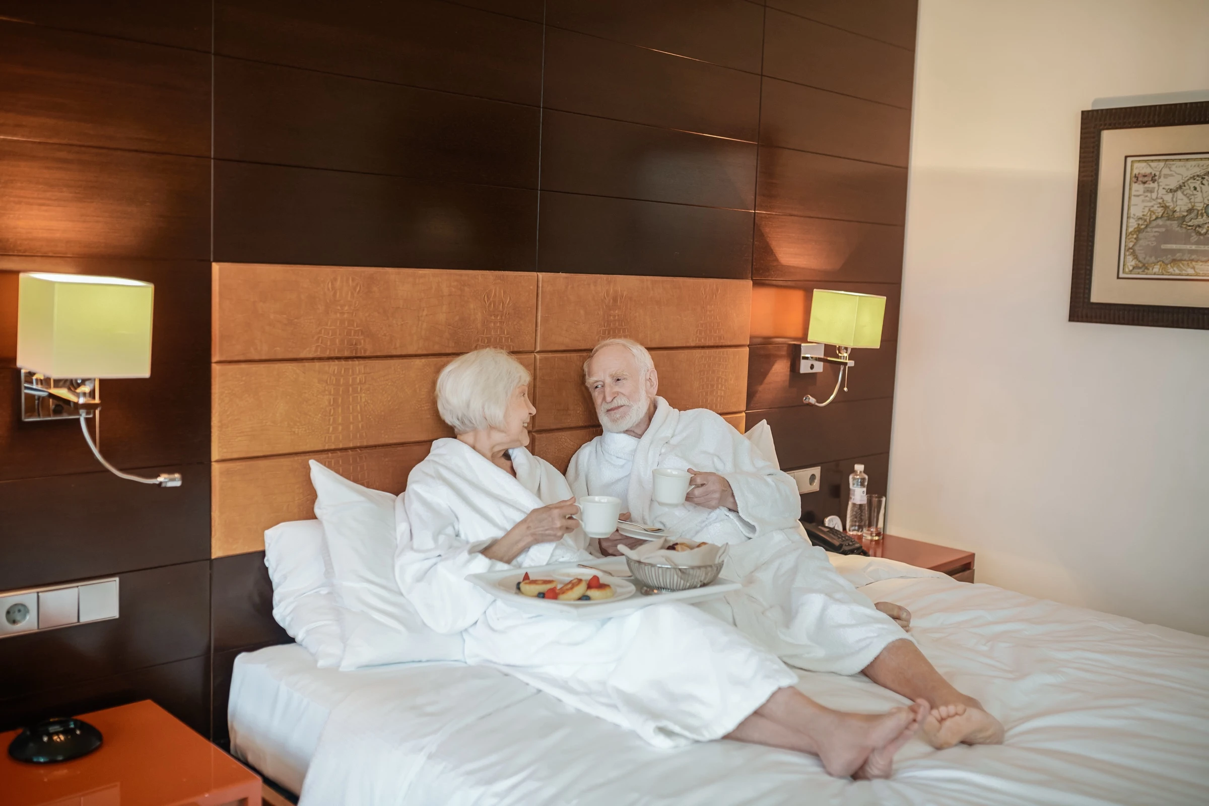 Couple senior souriant et détendu dans une chambre EHPAD haut de gamme spacieuse et confortable, ambiance hôtellière chaleureuse et lumineuse
