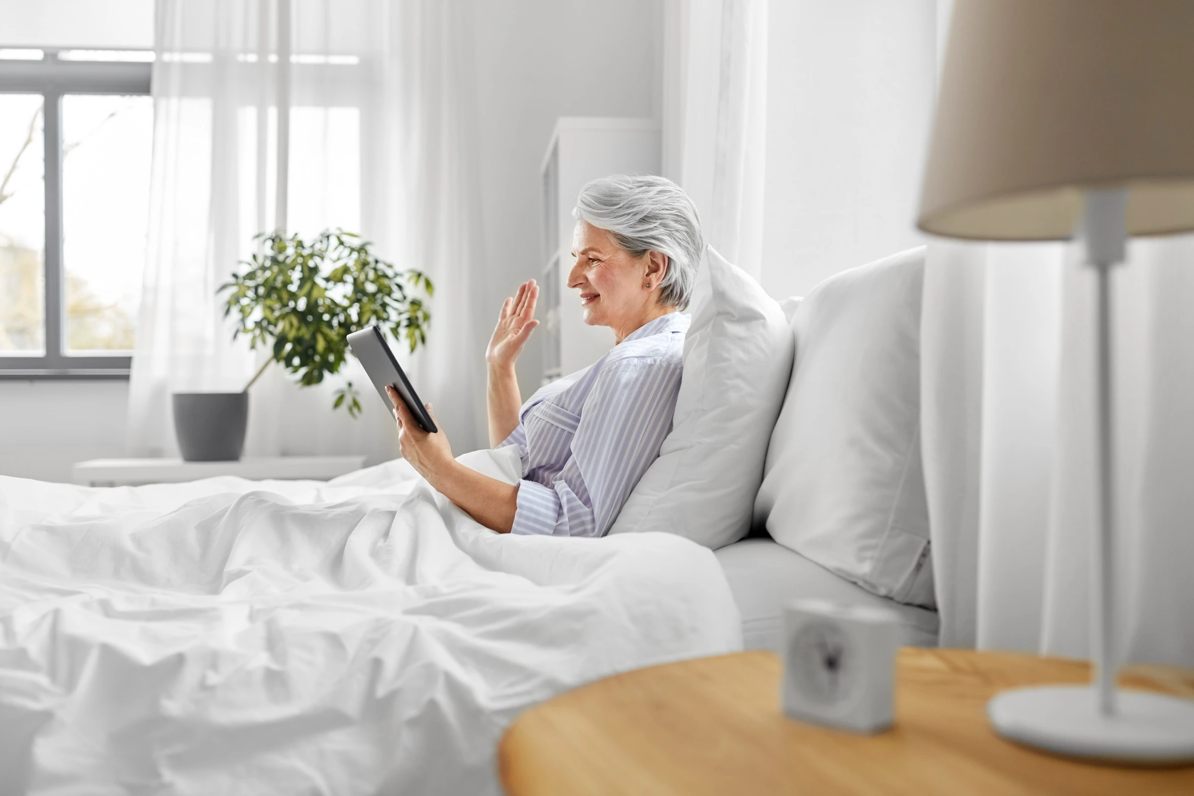Femme âgée dans sa chambre EHPAD souriant en visio avec sa famille sur une tablette numérique, lien préservé et chambre lumineuse personnalisée