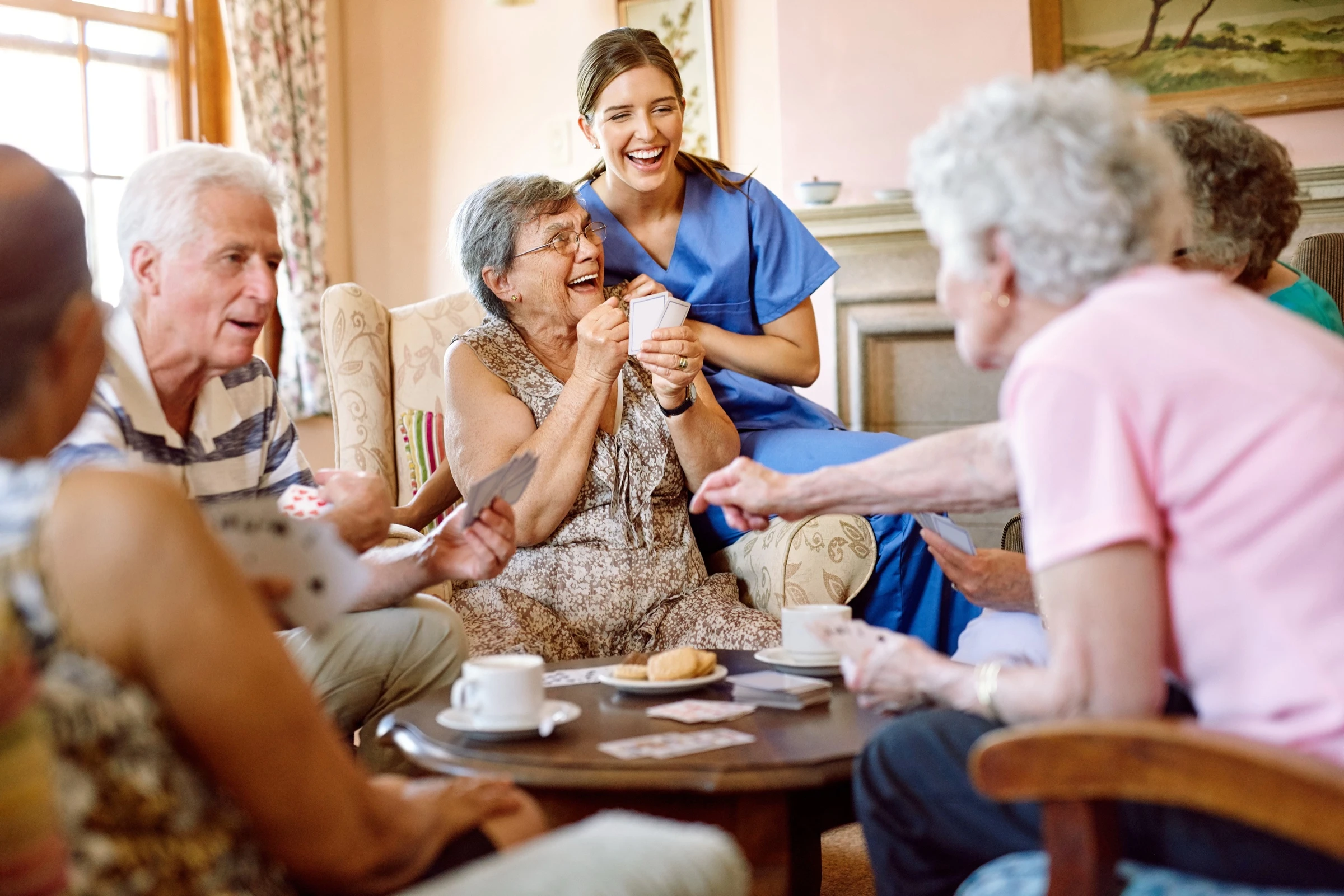 Groupe de résidents seniors jouent aux cartes en EHPAD avec une soignante qui les observe, équipe pluridisciplinaire et lien intergénérationnel