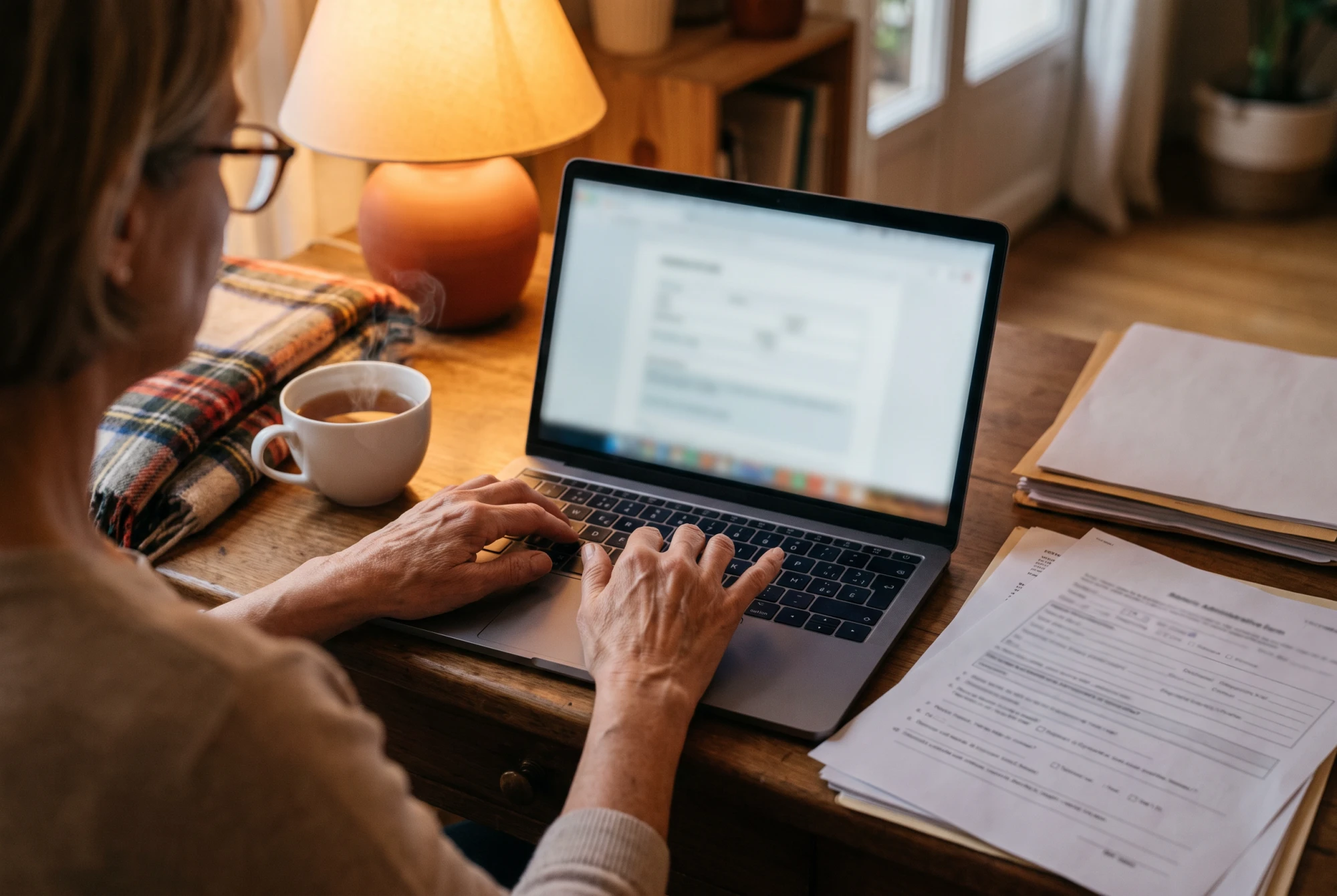 Mains d'une aidante remplissant un dossier administratif d'employeur sur un ordinateur portable, tasse de thé à côté, le soir.