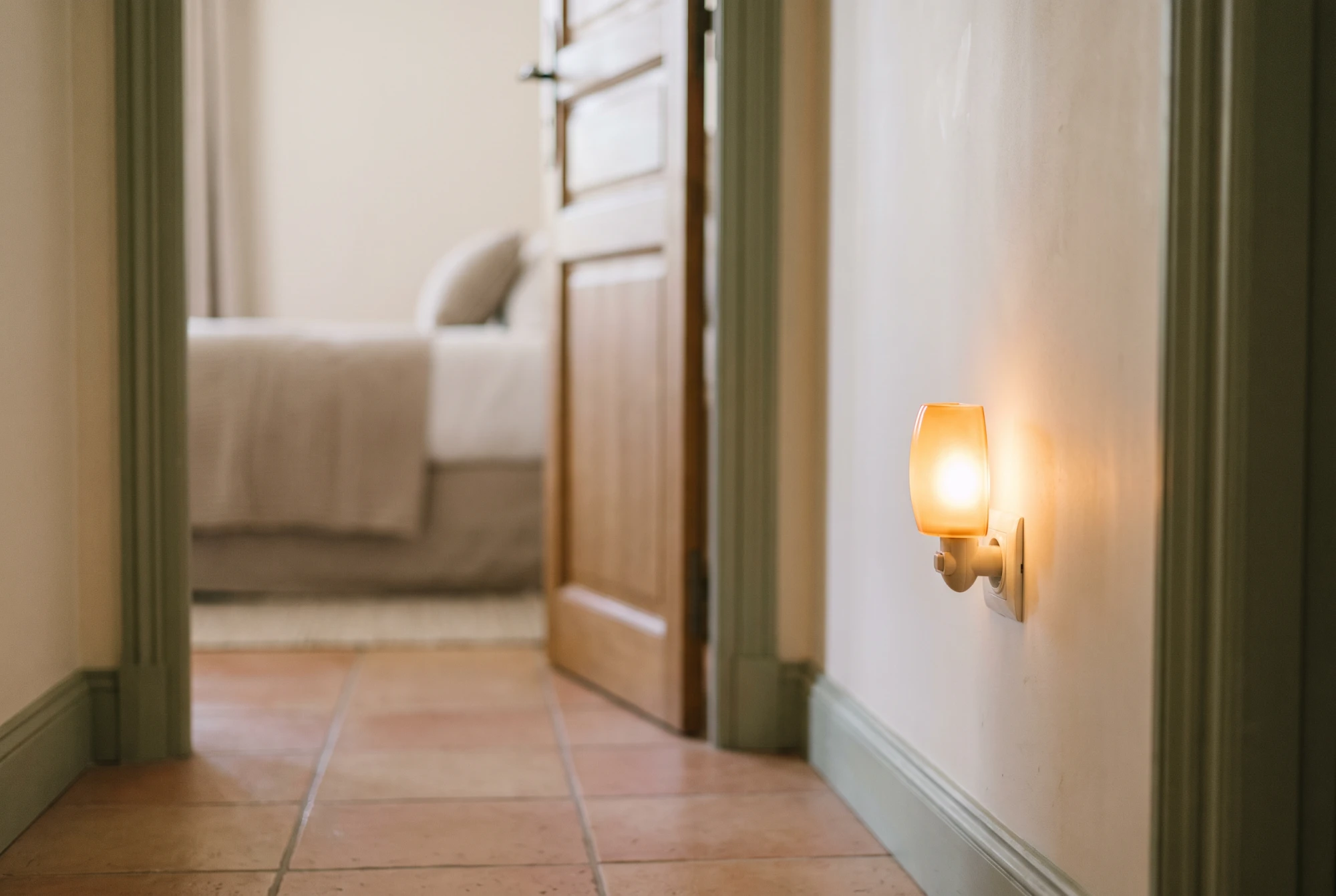 Veilleuse chaude allumée dans un couloir la nuit, porte de chambre entrouverte sur un intérieur paisible aux tons crème et terracotta.