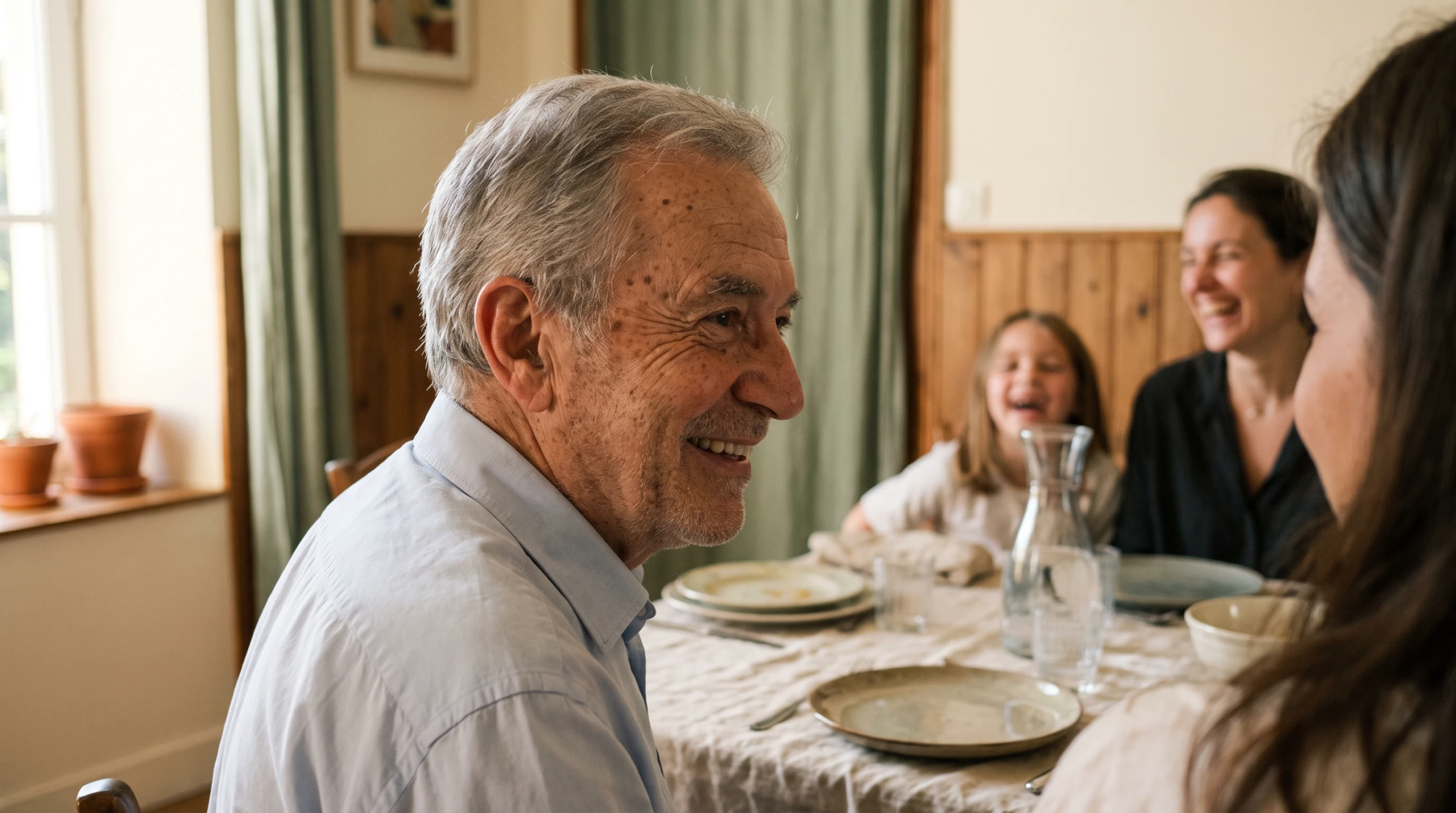 Homme de 72 ans souriant dans un repas de famille, il suit la conversation grâce à son appareil auditif invisible.