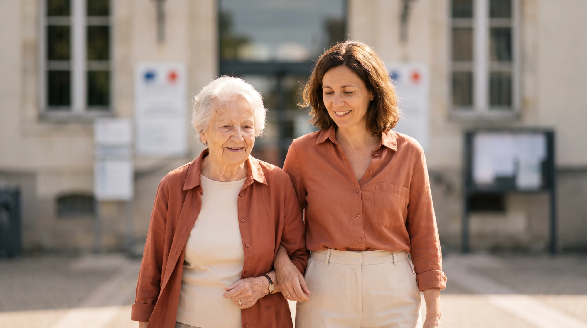 Femme âgée et sa fille aidante marchant ensemble devant la façade d'un bâtiment administratif départemental