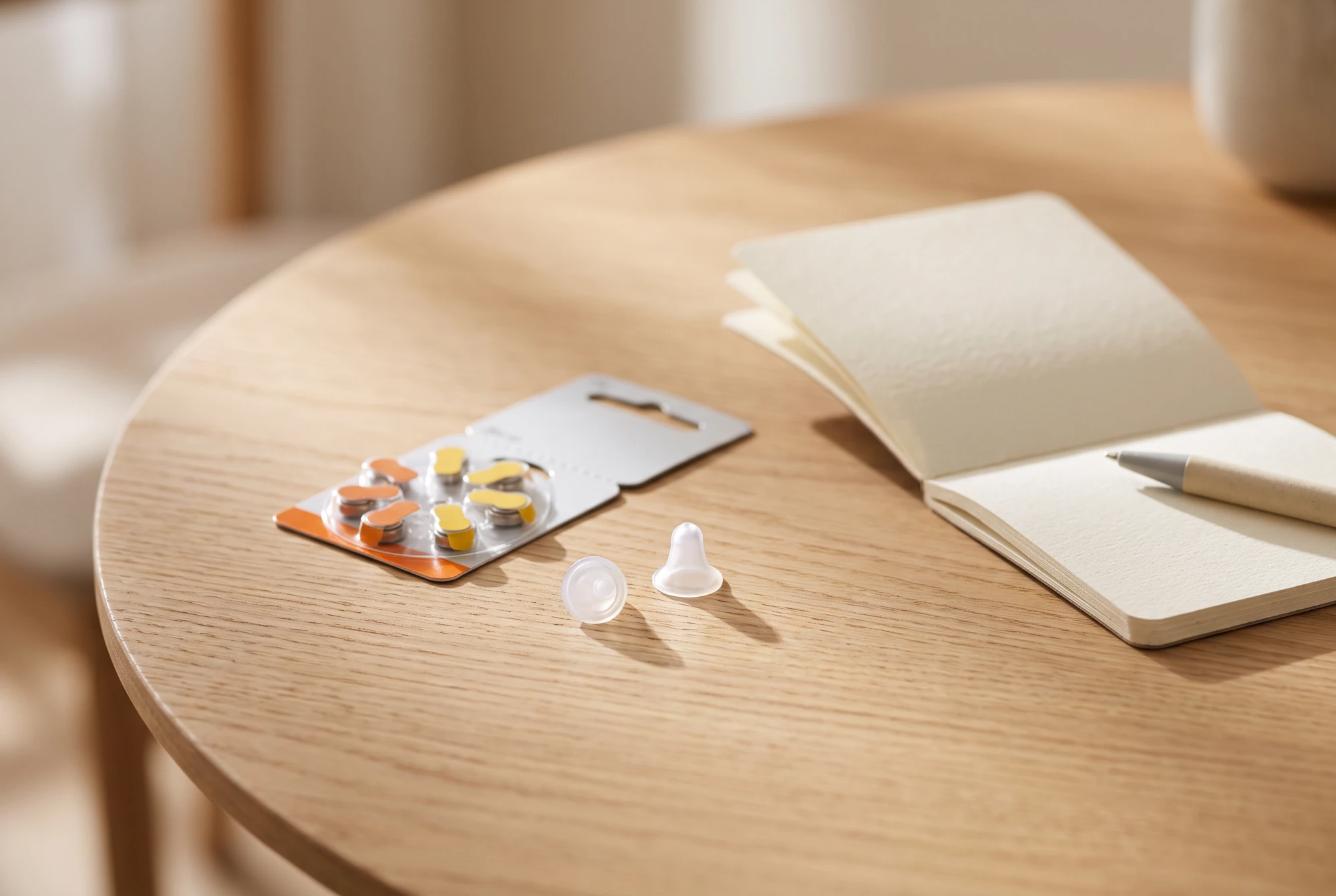Plaquette de piles auditives zinc-air et dôme en silicone posés près d'un carnet de budget sur une table en bois clair.