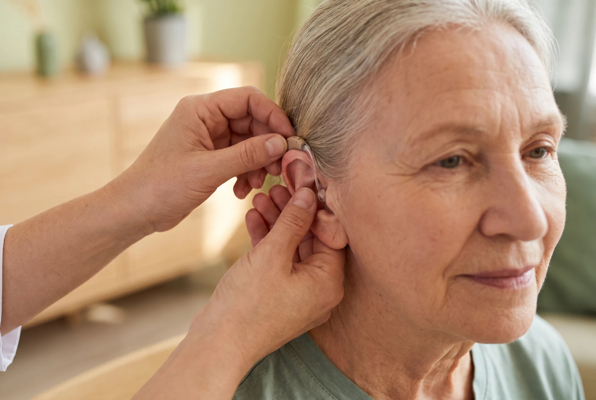 Audioprothésiste réglant avec soin un appareil auditif discret derrière l'oreille d'une personne âgée sereine, en cabinet clair.
