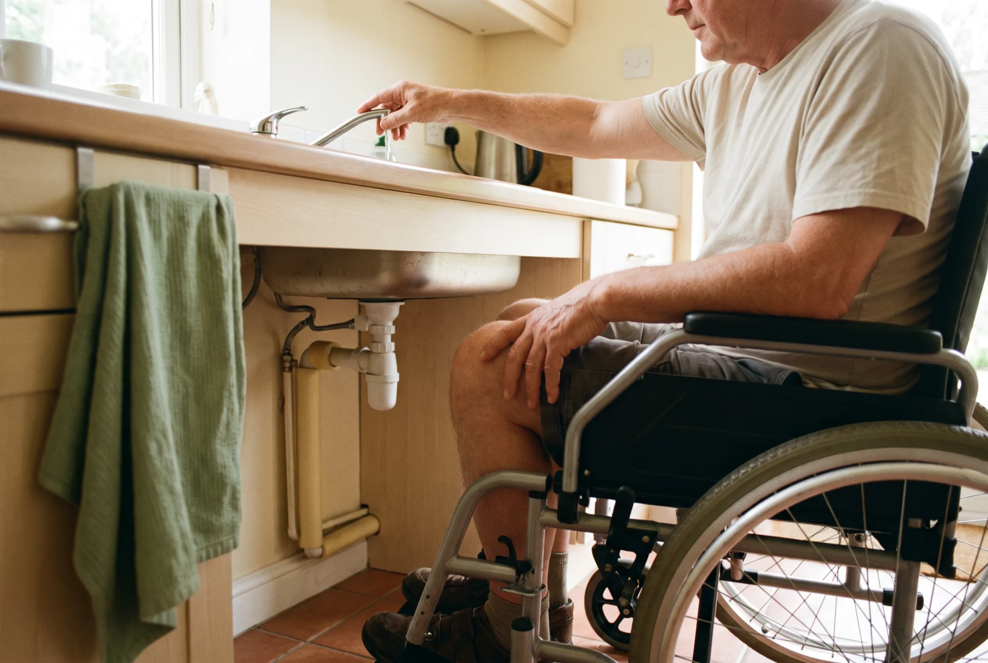 Homme en fauteuil roulant glisse ses genoux sous un évier accessible et actionne le mitigeur latéral.
