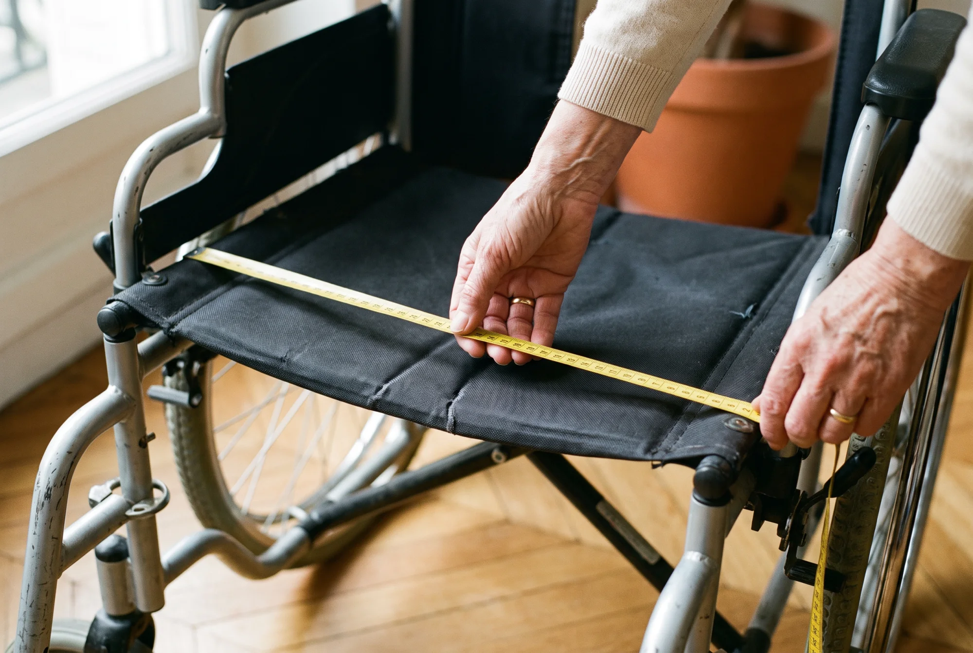 Mains d'une aidante mesurant au mètre ruban la largeur d'assise d'un fauteuil roulant d'occasion pliable.